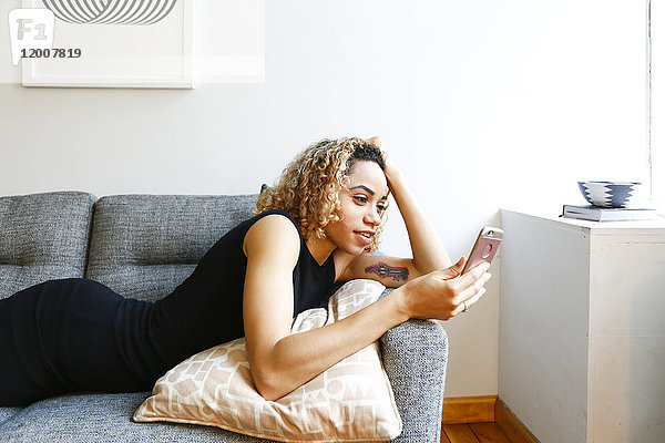 Gemischtrassige Frau liegt auf dem Sofa und schreibt eine SMS auf ihrem Handy