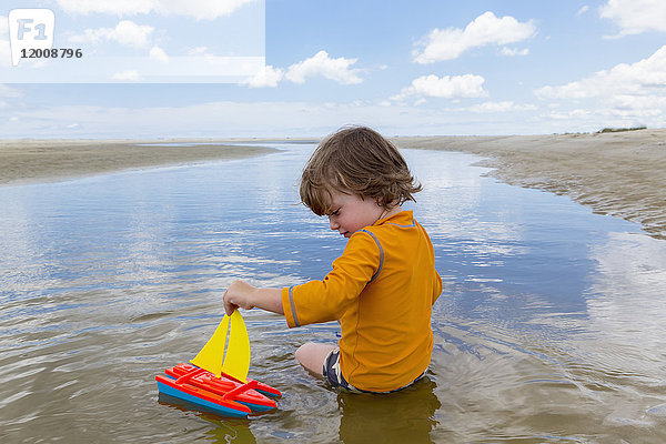 Kaukasischer Junge sitzt im Wasser und spielt mit einem Spielzeug-Segelboot