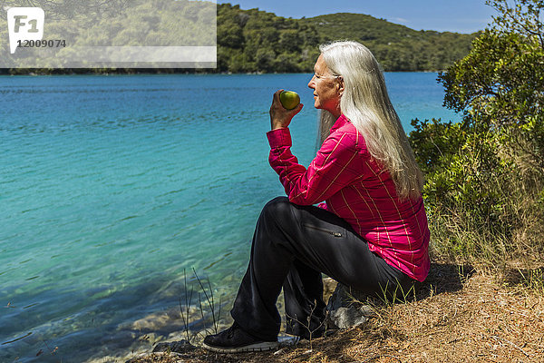 Kaukasische Frau sitzt am See und isst einen Apfel