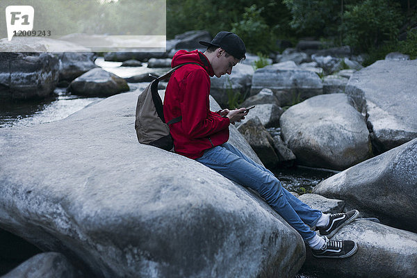Kaukasischer Mann sitzt auf einem Felsen im Fluss und schreibt eine SMS mit seinem Handy