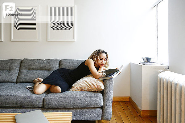Gemischtrassige Frau liegt auf dem Sofa und liest ein Buch