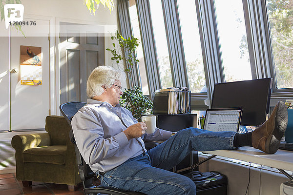 Kaukasischer Mann  der Kaffee trinkt und einen Laptop benutzt und die Füße auf den Schreibtisch legt