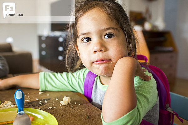 Unordentliches gemischtrassiges Mädchen isst Essen und trägt Rucksack am Tisch