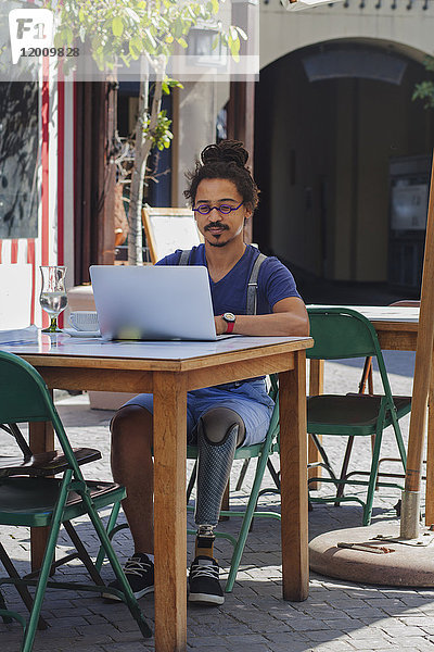 Gemischtrassiger Mann mit Beinprothese benutzt Laptop in einem Straßencafé