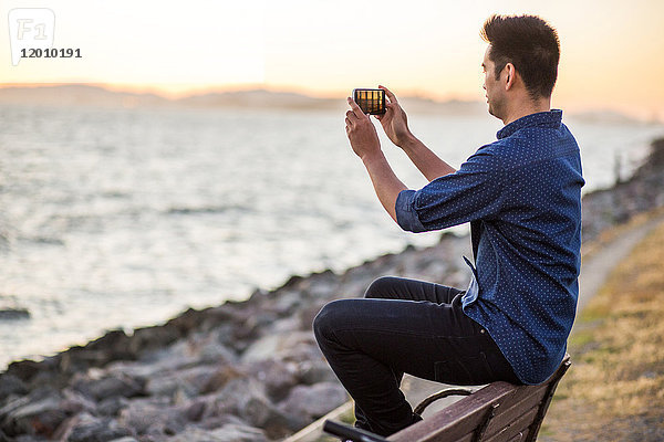 Chinesischer Mann sitzt auf einer Bank und fotografiert den Ozean mit seinem Mobiltelefon