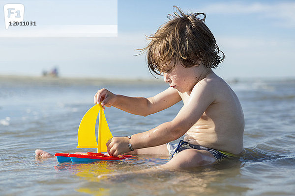 Kaukasischer Junge sitzt im Meer und spielt mit einem Spielzeug-Segelboot