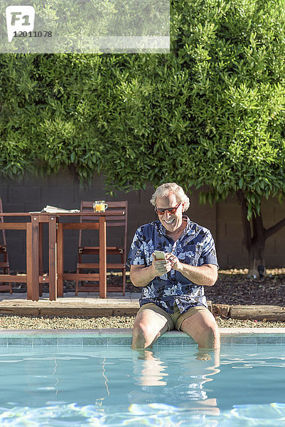 Kaukasischer Mann  der mit den Beinen im Schwimmbad eine SMS auf dem Handy schreibt