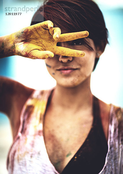 Junge Frau hält zwei mit Farbe befleckte Finger vor ihr Gesicht und schaut in die Kamera. Die Farbe bedeckte Kleidung und Haut.