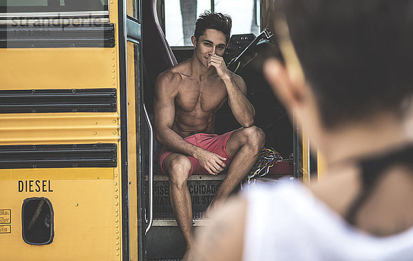 Junger Mann sitzt auf den Stufen eines Schulbusses und sieht eine junge Frau an.