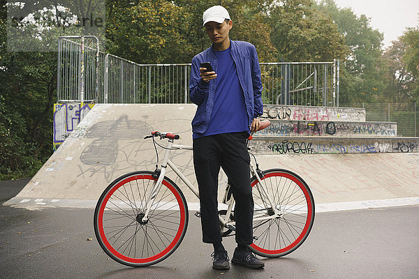 Volle Länge des Mannes mit dem Handy beim Stehen mit dem Fahrrad im Park