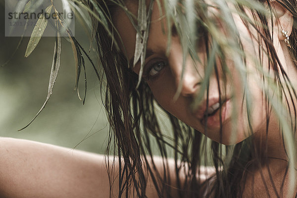 Nahaufnahme einer jungen Frau mit nassen Haaren  die durch die Blätter schaut.