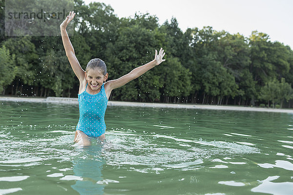 Fröhliches Mädchen mit erhobenen Armen  das im Schwimmbad gegen Bäume genießt.