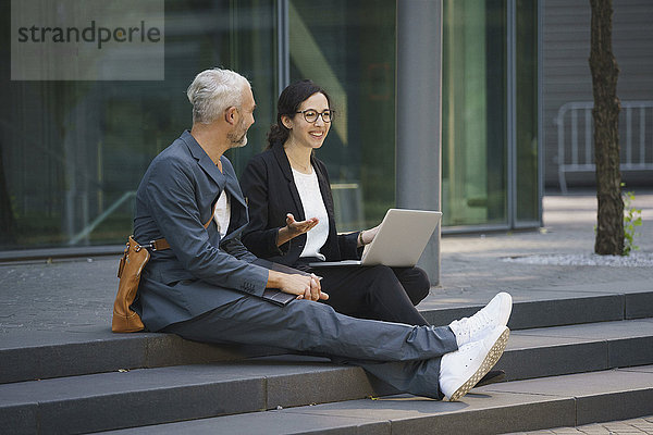 Lächelnde Geschäftskollegen mit Laptop diskutieren auf Stufen in der Stadt