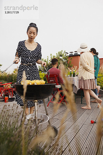 Junge Frau beim Grillen auf der Terrasse mit Freunden im Hintergrund