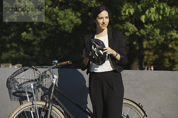 Lächelnde Geschäftsfrau  die an einem sonnigen Tag den Helm am Fahrrad gegen die Pflanzen hält.