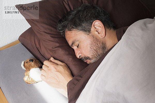 Hochwinkelansicht des erwachsenen Mannes  der von einem kleinen Teddybären auf dem Bett zu Hause schläft.