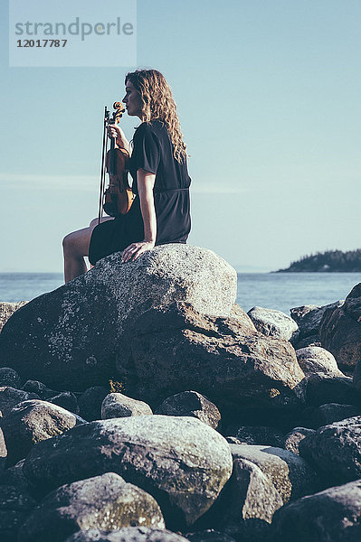 Flachwinkelansicht einer Frau mit Geige  die auf Felsen am See sitzt und dabei wegschaut