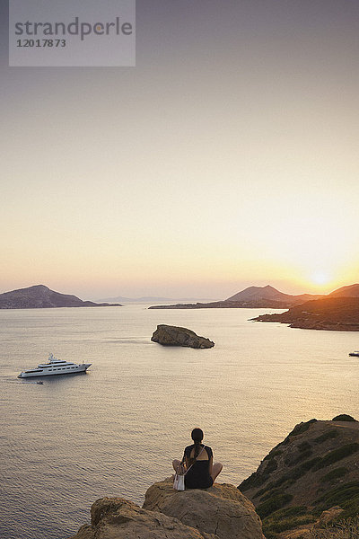 Rückansicht einer Frau  die auf einer Felsformation sitzt und bei Sonnenuntergang das Meer gegen den Himmel betrachtet.