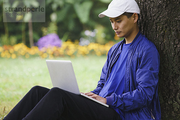Junger Mann mit Laptop beim Sitzen am Baum im Park