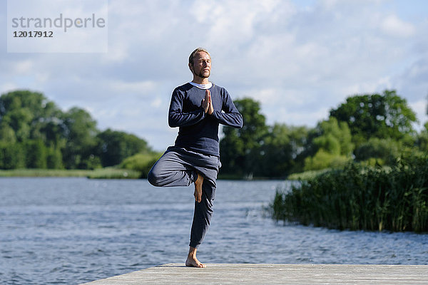 Reifer Mann praktiziert Baum-Pose Yoga auf Pier am See gegen den Himmel