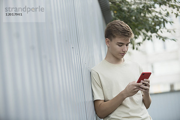 Neigungsbild eines Teenagers  der sein Handy benutzt  während er sich an eine Metallwand lehnt.