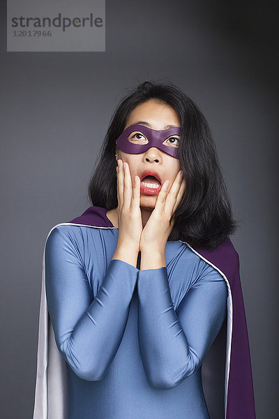 Überraschte Frau in Superheldenkostüm stehend mit Kopf in Händen vor grauem Hintergrund