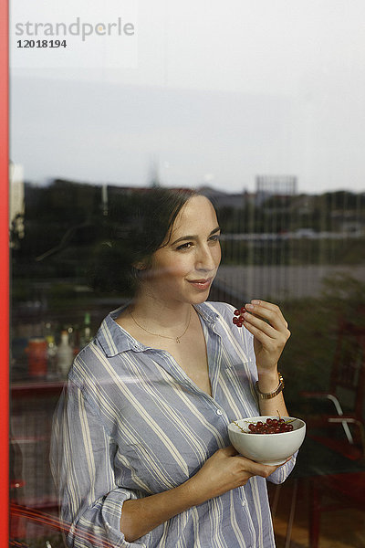 Nachdenkliche junge Frau isst rote Johannisbeeren  während sie durchs Fenster schaut.