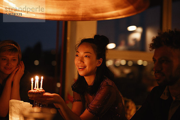 Glückliche Frau hält kleinen Geburtstagskuchen mit angezündeten Kerzen  während sie bei Freunden zu Hause sitzt.