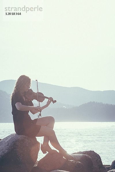 Frau spielt Geige  während sie auf dem Felsen am Seeufer gegen den klaren Himmel sitzt.