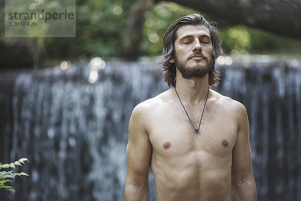 Hemdenloser junger Mann mit geschlossenen Augen  der am Wasserfall im Wald steht