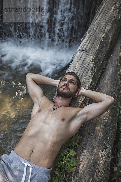 Nachdenklicher Mann ohne Hemd  der auf Baumstämmen über dem Wasserfall im Wald liegt.