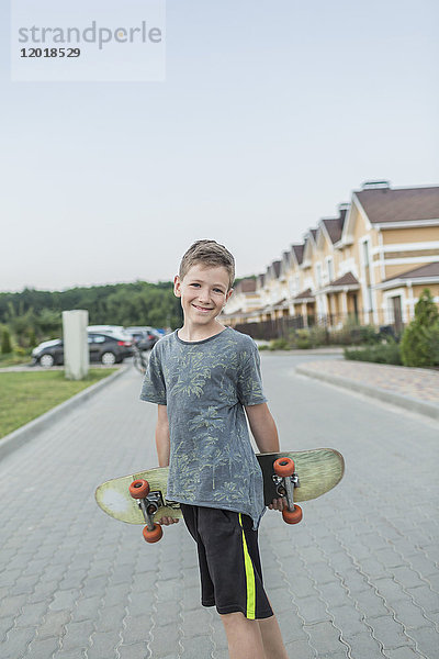 Porträt eines lächelnden Jungen  der Skateboard hält  während er auf der Straße gegen den klaren Himmel steht.
