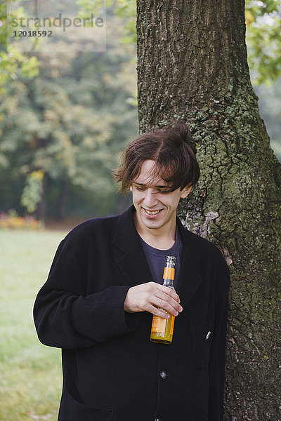 Junger Mann trinkt Saft  während er am Baum steht.