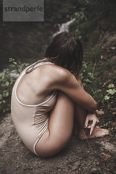 Sinnliche junge Frau in Badebekleidung auf einem Felsen im Wald sitzend