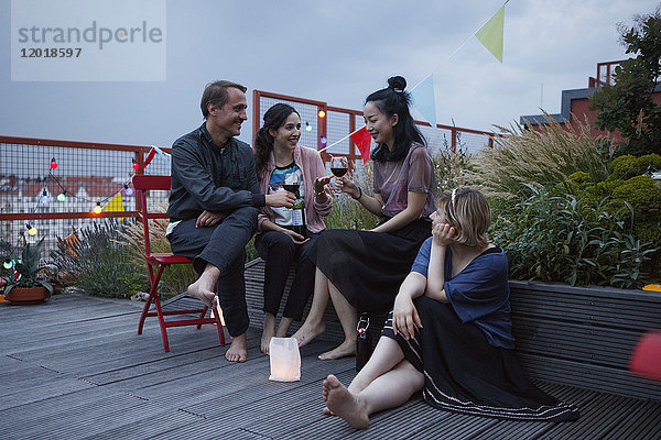 Freunde unterhalten sich  während sie in der Abenddämmerung auf der Terrasse Weingläser halten.