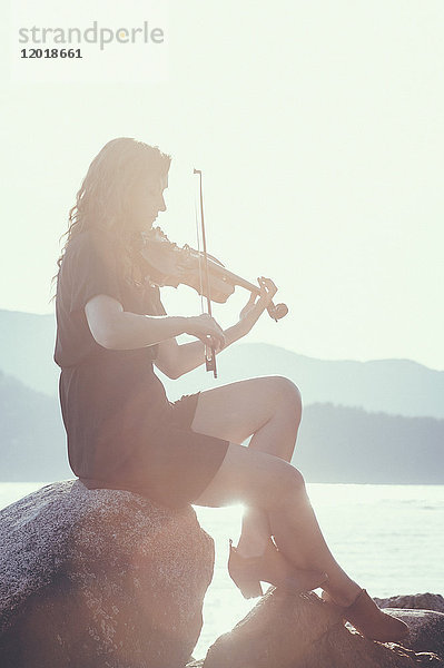 Volle Länge der hinterleuchteten Frau beim Geigenspiel auf dem Felsen am Seeufer gegen den klaren Himmel.