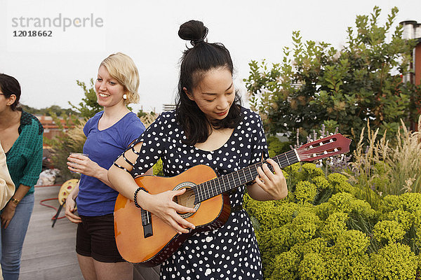 Junge Frau spielt Gitarre auf der Terrasse  während weibliche Freunde im Hintergrund stehen.