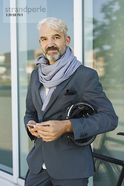 Porträt eines selbstbewussten Geschäftsmannes mit Handy und Fahrradhelm im Stehen gegen Glas