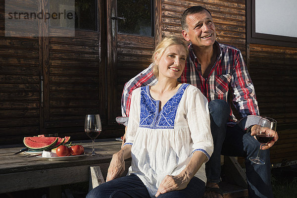 Lächelndes reifes Paar mit Wein und Wassermelone beim Entspannen vor dem Bauernhaus