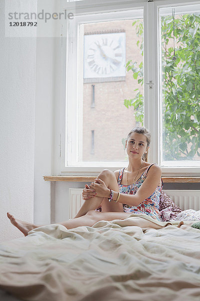 Schöne junge Frau sitzt zu Hause auf dem Bett gegen das Fenster.