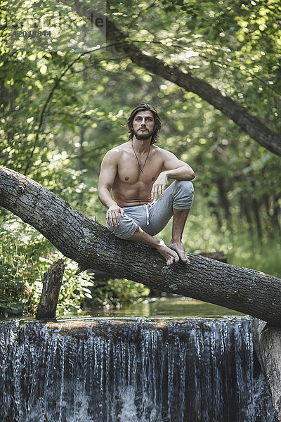 Nachdenklicher Mann ohne Hemd sitzt auf einem Ast über einem Wasserfall im Wald.