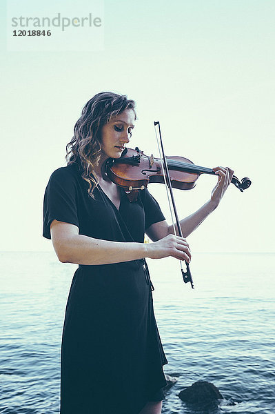 Schöne junge Frau spielt Geige  während sie am See gegen den klaren Himmel steht.