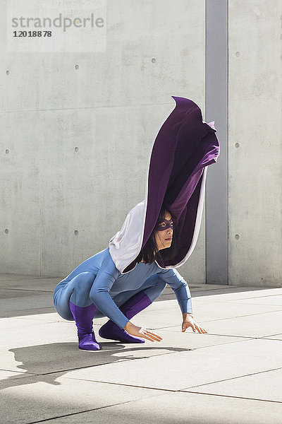 Weibliche Superheldin mit violettem Umhang auf dem Boden an der Wand kauernd