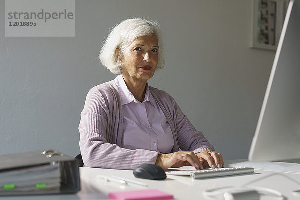Porträt einer älteren Frau  die im Büro an der Wand sitzt und den Computer benutzt.
