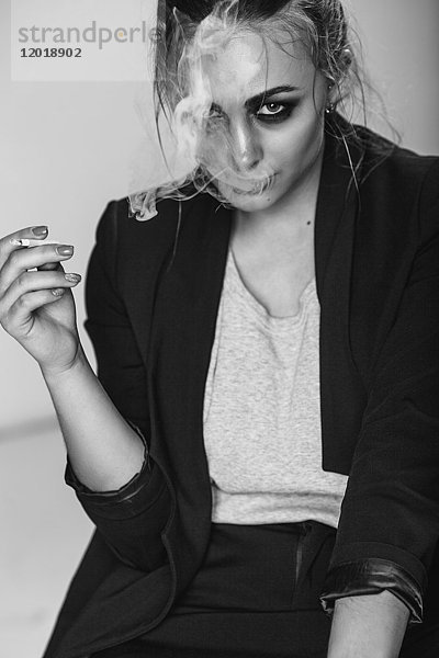 Porträt einer jungen Frau  die im Sitzen vor grauem Hintergrund eine Zigarette raucht.