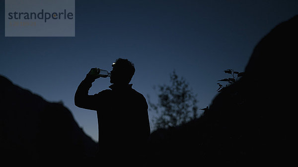 Silhouette Mann trinkt Wasser aus der Flasche  während er nachts auf dem Berg steht.