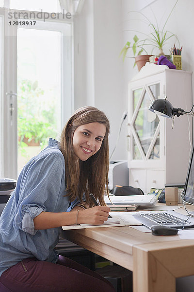 Porträt einer lächelnden jungen Frau beim Schreiben am Computertisch im Zimmer
