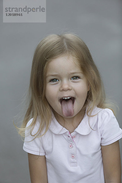Portrait eines fröhlichen Mädchens  das die Zunge vor grauem Hintergrund herausstreckt.