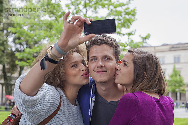 Frau nimmt Selfie mit einer Freundin  während sie einen jungen Mann küsst.