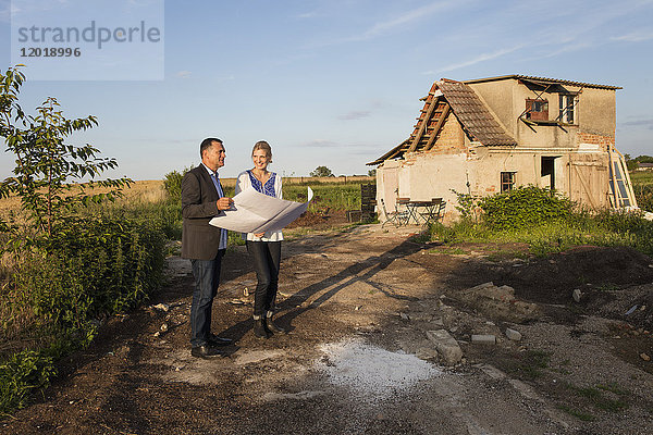 Lächelnde Frau mit Immobilienmaklerin beim Lesen des Dokuments  während sie sich gegen das Bauernhaus stellt.